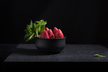 Fresas enteras con hojas de menta en un recipiente negro sobre una mesa de madera negra y fondo oscuro © R. Guas