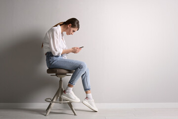 Woman with bad posture using smartphone while sitting on stool near light grey wall indoors, space for text - obrazy, fototapety, plakaty