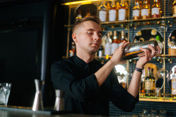 Focused professional barman mixing ingredients of alcoholic cocktail by shaking shaker standing behind bar counter, on blurred background of shelves with different alcoholic drinks, selective focus.
