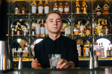 Portrait of young serious bartender standing behind bar counter and holding stirring spoon and glass filled with ice cubes, preparing to make cocktail, on background of shelves with alcoholic drinks.