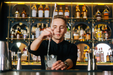 Portrait of bartender male stirring ice cubes in glass using spoon standing on bar counter, on background of shelves with different alcoholic drinks. Barman making cocktail in bar with dark interior.
