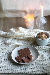 Plate with almond chocolate bar, mug with chocolate chip cookies, open book, reading glasses, soft blanket and lit candles. Hygge at home. Selective focus.