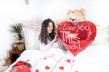 A beautiful woman in pink pajamas with a heart-shaped balloon lies on bed at home.