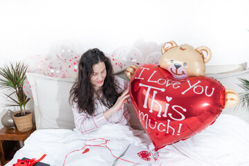A beautiful woman in pink pajamas with a heart-shaped balloon lies on bed at home.