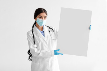 Serious millennial indian woman therapist in white coat with stethoscope in protective mask, gloves hold banner