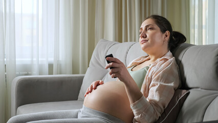 Bored pregnant woman with brunette hair in ponytail wearing flannel and sports top switches TV...