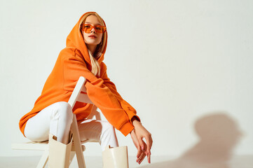 Young confident blonde girl wearing trendy orange hoodie, color sunglasses, posing on white background. Studio fashion portrait. Copy, empty space for text - obrazy, fototapety, plakaty