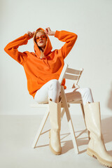 Fashionable confident woman wearing trendy orange hoodie, sunglasses, white skinny jeans, high boots, sitting, posing on chair. Full body portrait - 484876193