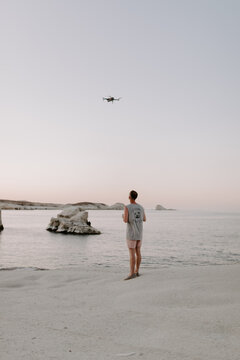 young male traveller is using drone to film and photograph aerial views of a beach in Greece, surrounded by rocky white cliffs and blue water at sunset