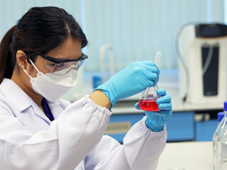 The scientist wears PPE and holds an Erlenmeyer flask laboratory, with red solvent solution from...