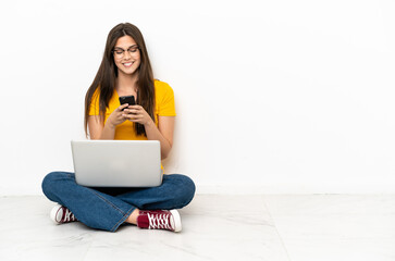 Young woman with a laptop sitting on the floor sending a message with the mobile