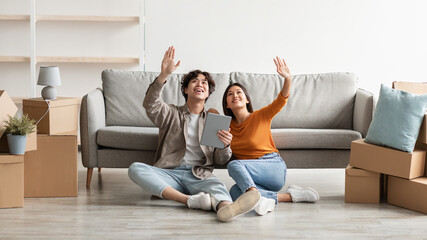 Millennial Asian couple sitting on floor among carton boxes, using digital tablet, dreaming about design of new home
