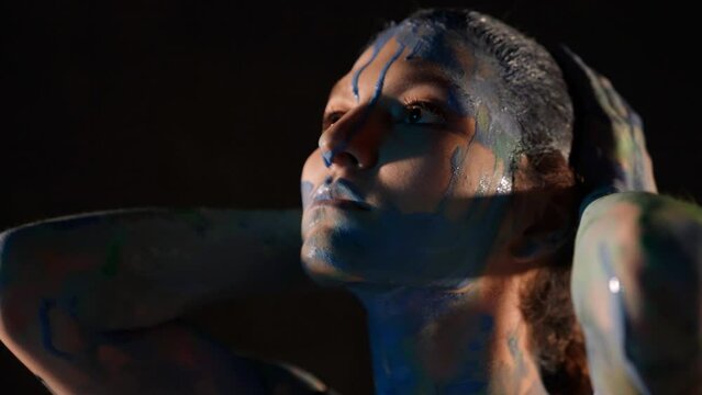 passionate lady with blue paints on skin is stroking her head, portrait in dark room
