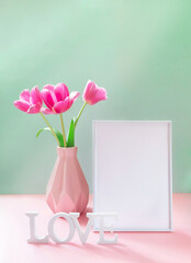 Pink tulips in vase, empty white frame, white letters LOVE on pastel pink green backdrop. Mock up.