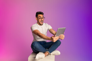 Emotional young Arab man sitting cross legged with digital tablet, looking at screen in shock, neon light