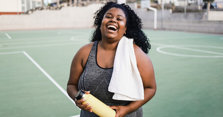 Plus size african woman smiling at camera while doing running routine in park city - Focus on face - obrazy, fototapety, plakaty