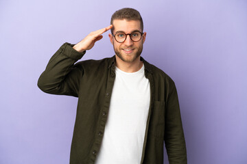 Young handsome caucasian man isolated on purple background saluting with hand with happy expression