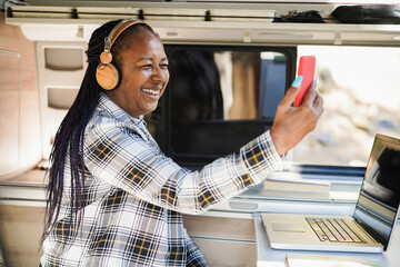 African senior woman having fun doing video call and wearing headphones inside camper mini van -...