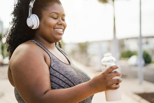 Curvy woman drinking while doing jogging routine outdoor at city park - Focus on headphones