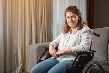 Young disabled woman with infantile cerebral paralysis in white shirt smiles sitting in wheelchair near stylish sofa in living room at home, sunlight. - obrazy, fototapety, plakaty