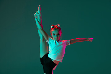 Little flexible girl, rhythmic gymnastics artist training isolated on green studio background in neon pink light. Grace in motion, action. Doing exercises in flexibility. - obrazy, fototapety, plakaty