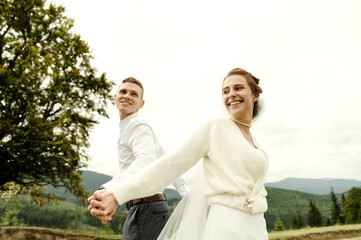 cheerful bride and groom stand with their backs to each other and hold hands on the top of the mountain. wedding day of a young couple in love