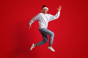 Holiday Fun. Cheerful Young Man Wearing Santa Hat Jumping Over Red Background