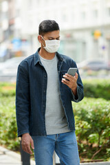 Serious young man in medical mask walking in the street and reading news on coronavirus on smartphone