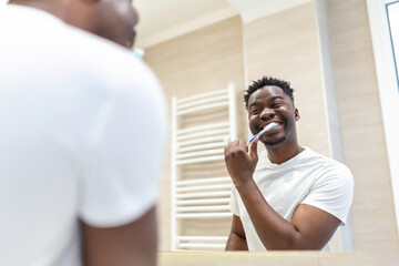Smiling African man with toothbrush cleaning teeth and looking mirror in the bathroom. Handsome young man brushing his teeth in morning in bathroom. brushing teeth at night before going to sleep.