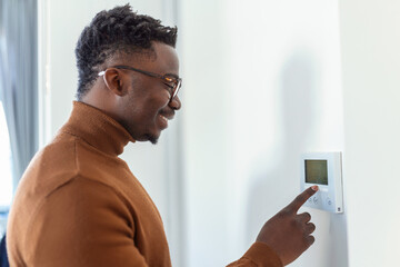 Smiling African American man using modern smart home system, controller on wall, positive young man switching temperature on thermostat or activating security alarm in apartment - obrazy, fototapety, plakaty