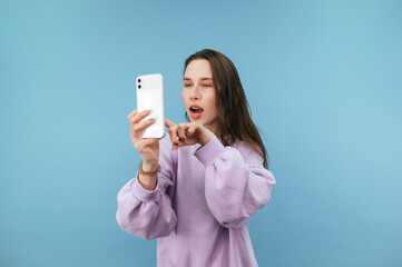 Cheerful woman in sweatshirt uses smartphone with surprised face on blue background, touches finger to phone screen