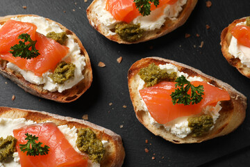 Delicious bruschettas with cream cheese, salmon and pesto sauce on black table, flat lay