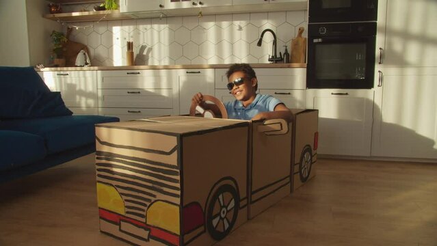 Cool carefree elementary age African boy in sunglasses driving toy cardboard car, holding steering wheel with one hand, expressing confidence and playful mood while playing in domestic room.