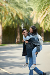 Happy young man giving piggyback ride to his girlfriend when they are crossing road in park