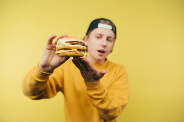 Young man in casual clothes and cap presents to the camera appetizing burger on a yellow background. Fast food concept