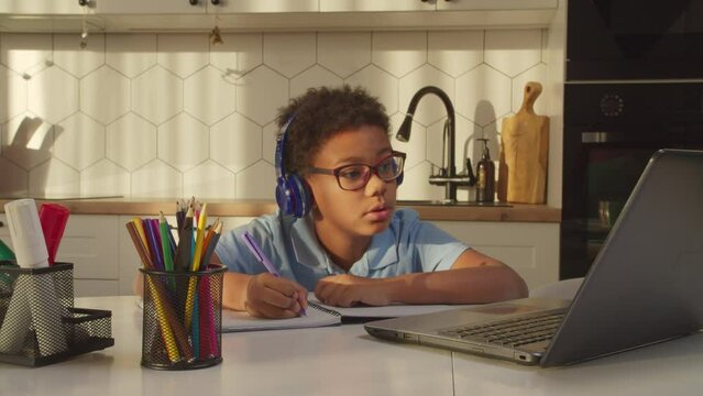 Smart cute black schoolboy in eyeglasses and wireless headphones studying online distance course using laptop pc, doing homework and writing assignment in notebook while learning at home.