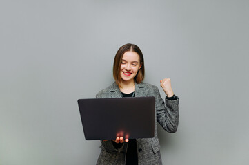 Joyful business young woman with a laptop in her hands rejoices with a smile on her face on a gray background.