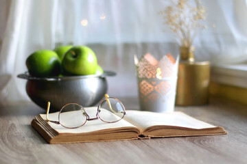 Bowl of green apples, open book, reading glasses, lit candle and flowers on the table. Selective focus.