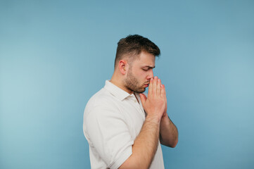 Adult man in a white T-shirt prays on a blue background with a serious face. A religious man with a bristle prays