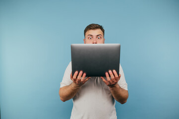 Portrait of shocked man with laptop in hands on blue background, looking at screen with funny emotional eyes.
