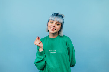 Cute teen girl with colored hair stands on a blue background and with a smile on his face shows his fingers gesture heart