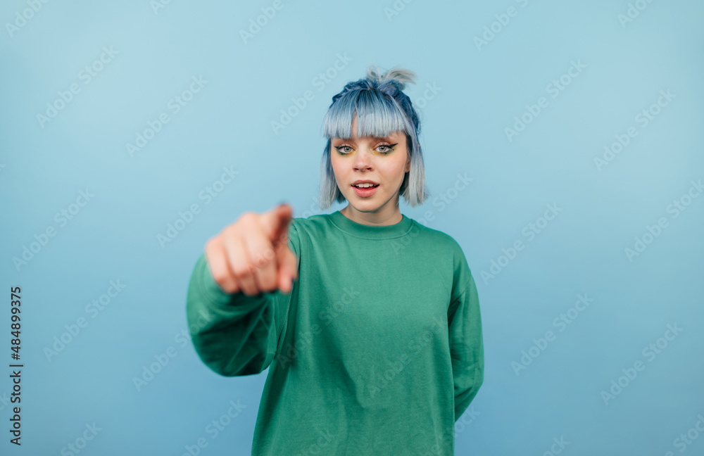 Canvas Prints Positive teen girl with blue hair points a finger at the camera and smiles on a blue background