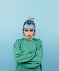 Serious girl with colored hair and in a green sweatshirt stands on a blue background and looks unhappily at the camera. Vertical
