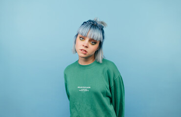 Portrait of a beautiful girl with blue hair and in a green sweatshirt on a blue background, posing for the camera with a serious face and biting her lip.