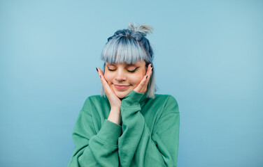 Beautiful lady in casual clothes and colored hair isolated on a blue background, shows a cute face holding her palms to her cheeks and smiling with her eyes closed.