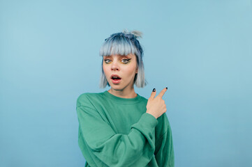 Surprised girl with blue hair looks at the camera with a shocked face and points his finger to the side, isolated on a blue background.