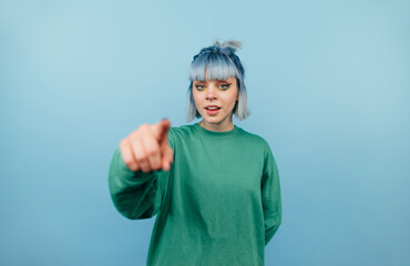 Positive teen girl with blue hair points a finger at the camera and smiles on a blue background
