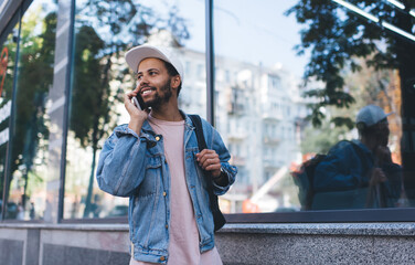 Hispanic male tourist calling to best friend for discussing hipster travelling during spring daytime, cheerful Latino man in cap connecting to 4g in roaming for making positive mobile communication