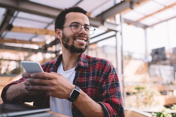Cheerful hipster guy with modern cellphone gadget in hand looking away and smiling during leisure time, happy millennial male blogger with digital mobile device enjoying youthful lifestyle