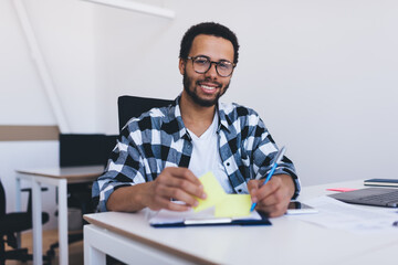 Portrait of happy male executive manager in eyeglasses smiling at camera while creating business plan and strategy for company, student posing at desktop enjoying time for exam